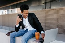 Smiling male entrepreneur with smart phone using laptop while sitting on retaining wall — Stock Photo