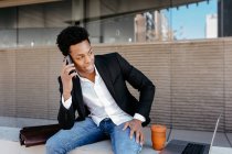 Усміхаючись чоловік - бізнесмен розмовляє на смартфоні сидячи на стіні і озираючись — стокове фото
