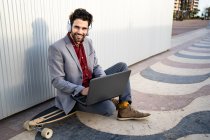 Hombre de negocios sonriente con portátil sentado en longboard frente a la pared - foto de stock