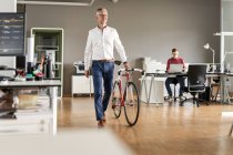 Mature businessman with bicycle walking in open plan office — Stock Photo