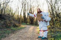 Female astronaut in space suit gesturing while holding cardboard on dirt road — Stock Photo