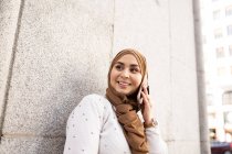 Junge Frau im Hidschab schaut weg, während sie mit dem Smartphone an der Wand spricht — Stockfoto