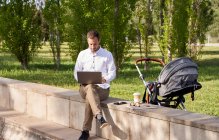 Mid adult businessman working on laptop while sitting by baby stroller in park — Stock Photo