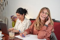 Mujer de negocios sonriente mirando hacia otro lado hablando por teléfono inteligente mientras está sentada con una colega en la cafetería - foto de stock