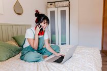 Young woman talking on mobile phone while using laptop in bedroom — Stock Photo