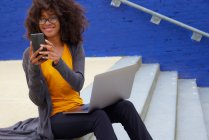 Souriant Afro femme en utilisant un téléphone portable tout en étant assis sur les marches — Photo de stock