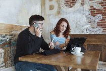 Hombre profesional hablando por teléfono inteligente mientras está sentado con una colega que trabaja en la cafetería - foto de stock