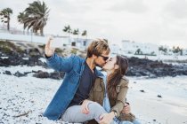 Парень целует девушку, делая селфи через смартфон на пляже — стоковое фото