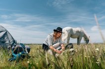 Young woman wearing hat sitting by horse grazing meadow during sunny day — Stock Photo