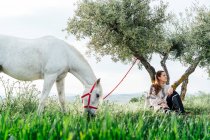 Young woman sitting in front of tree while horse grazing on field — Stock Photo