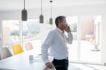 Empresario mirando hacia otro lado mientras habla por teléfono inteligente en la cocina doméstica en casa - foto de stock