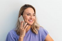 Lächelnde junge Frau schaut weg, während sie vor der Wand mit dem Handy telefoniert — Stockfoto