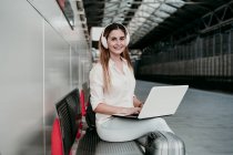 Glückliche Passagierin hört Musik über Kopfhörer, während sie mit Laptop am Bahnhof sitzt — Stockfoto