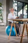 Smiling businesswoman working on laptop while sitting on fitness ball at desk in home office — Stock Photo