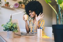 Afro woman with coffee cup waving during video call through laptop at home — Stock Photo