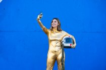 Woman in astronaut costume taking selfie through smart phone in front of blue wall — Stock Photo
