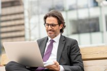 Hombre de negocios satisfecho con anteojos usando portátil mientras está sentado en el banco - foto de stock