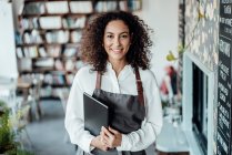 Smiling woman wearing apron holding book while standing at cafe — Stock Photo