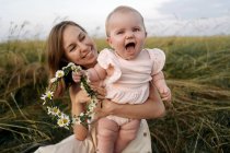 Sorrindo mãe segurando alegre bebê menina com flor tiara no campo — Fotografia de Stock