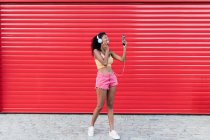 Smiling young woman taking selfie through smart phone while listening music in front of red shutter — Stock Photo