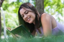 Sorridente giovane donna che legge il libro mentre giace sull'erba nel parco — Foto stock