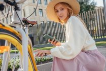 Jeune femme blonde tenant un téléphone intelligent tout en s'accroupissant à vélo — Photo de stock