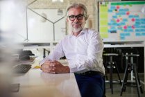 Mature businessman wearing eyeglasses sitting with hands clasped at desk in office — Stock Photo