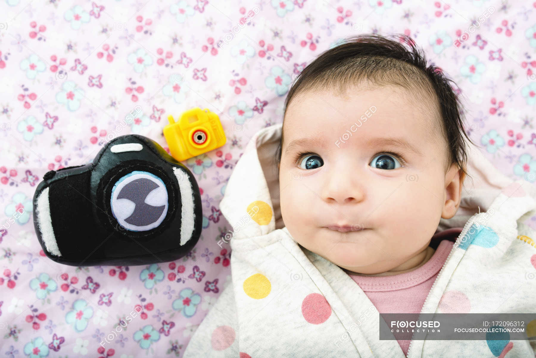 portrait-of-baby-girl-lying-on-bed-with-two-toy-cameras-toys-indoor