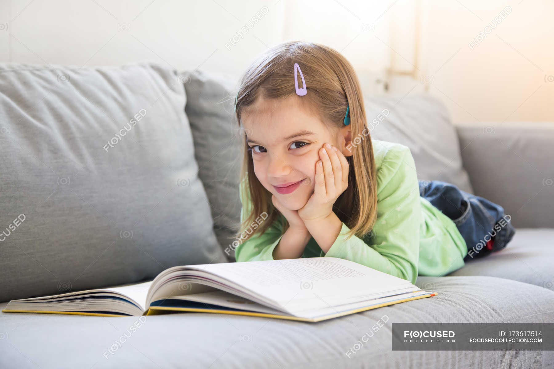 Portrait of smiling little girl lying on couch with book — female ...