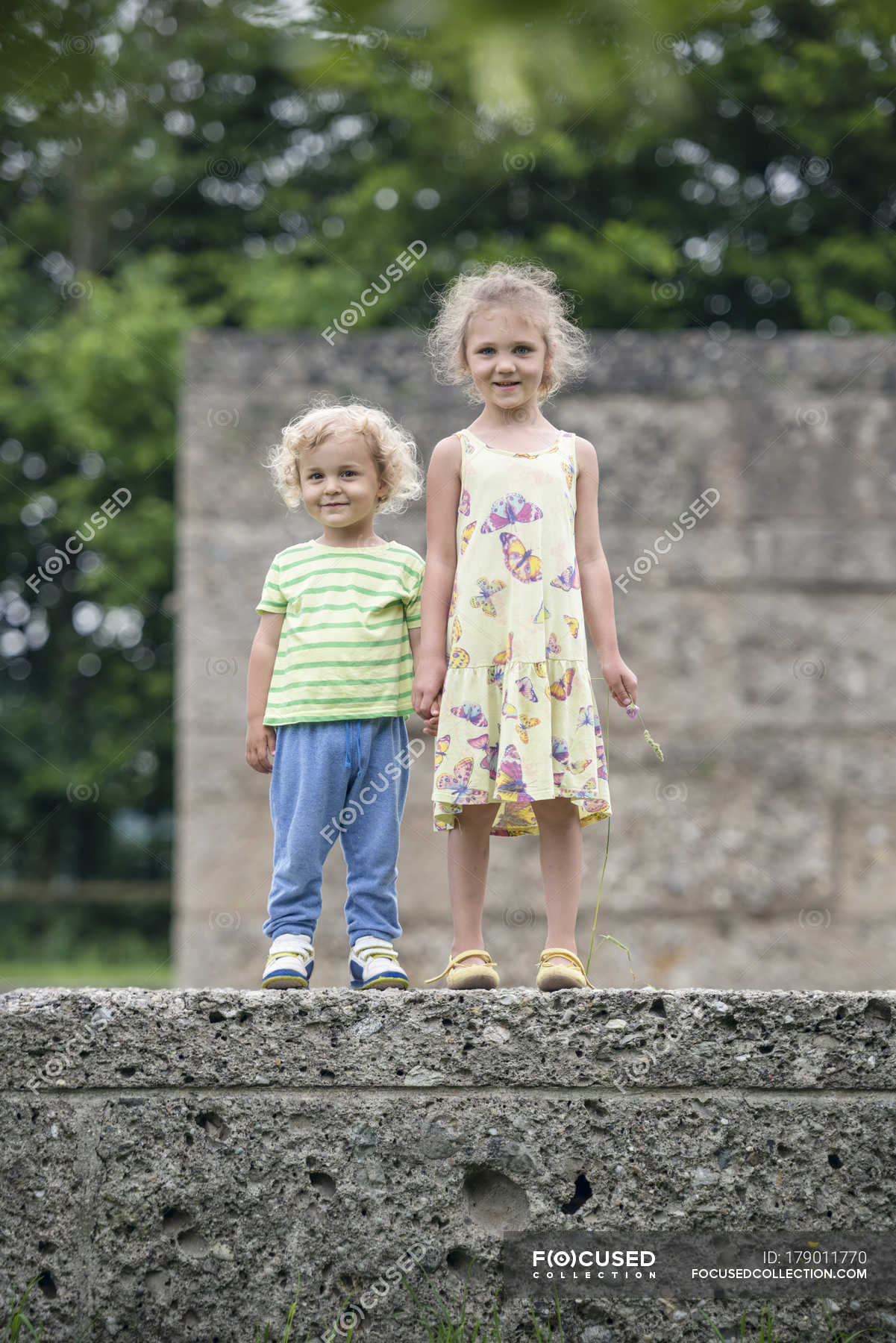 Portrait Of Little Sister And Brother Standing Side By Side On A Wall Holding Hands — People