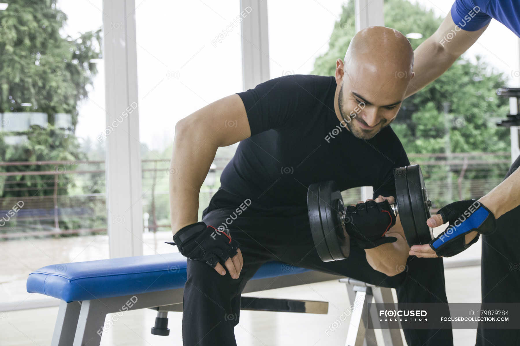 Man Lifting A Dumbbell Sitting On A Bench In A Gym Assisted By A