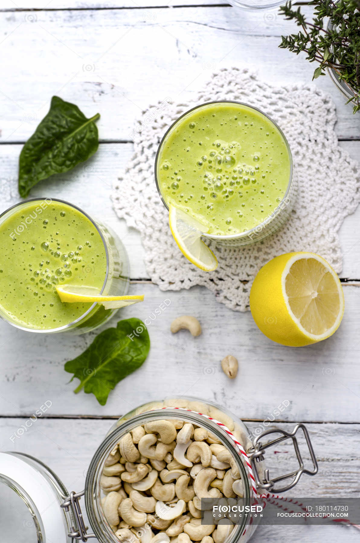 Two glasses of spinach smoothie, cashew nuts and half of lemon on white  wood — doily, ingredients - Stock Photo | #180114730