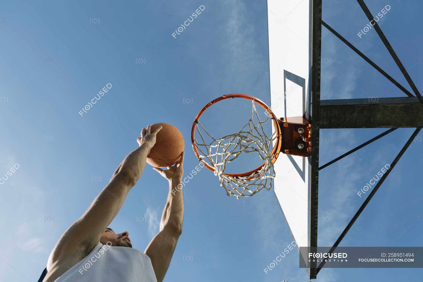 Ângulo baixo do jogador de streetball masculino mirando com bola enquanto  jogava basquete contra o céu azul