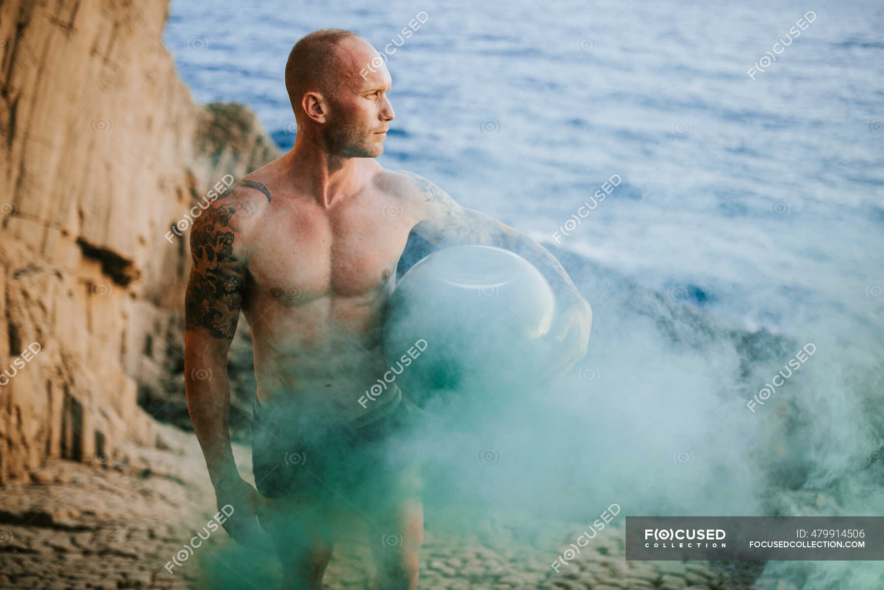 Tattooed Nudist Standing On Volcanic Rocks By The Sea Holding Fisghbowl With Colorful Smoke