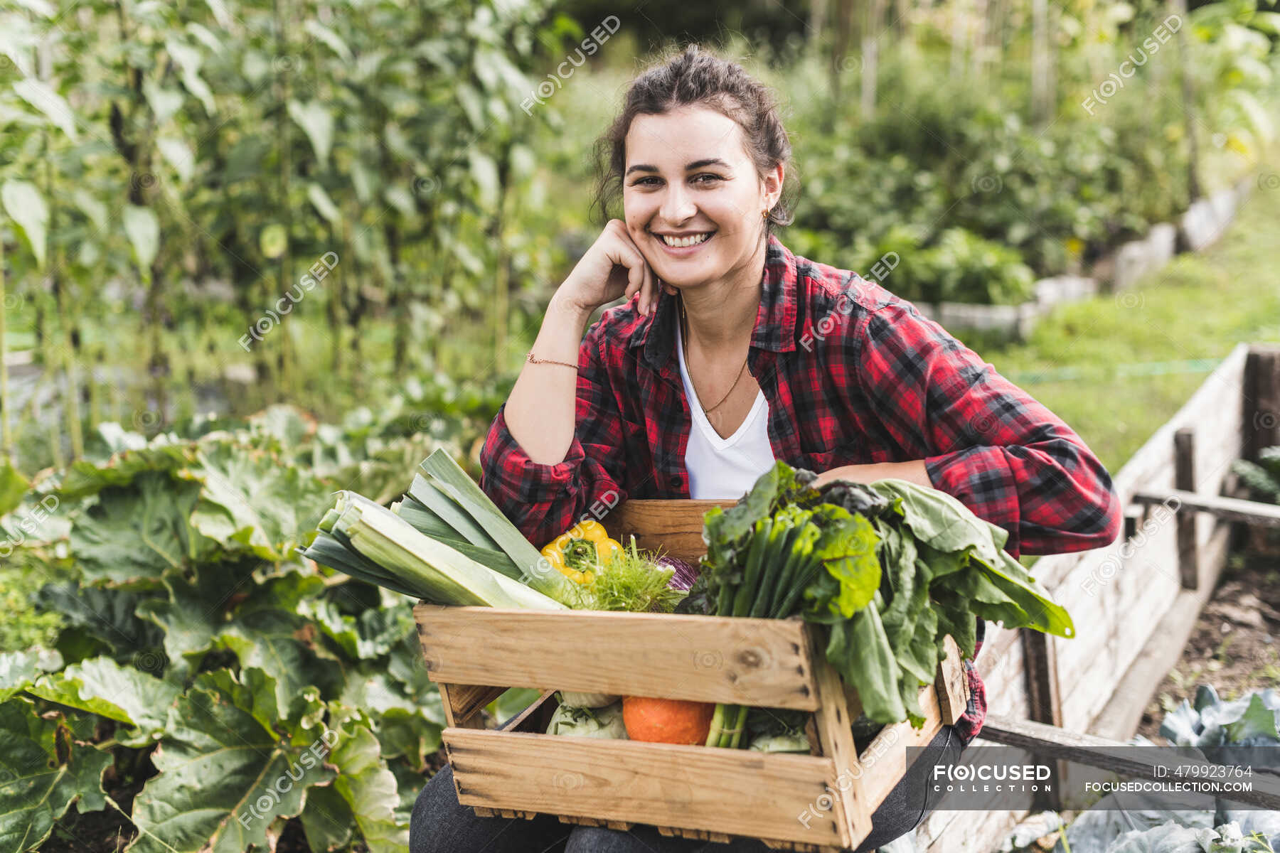 Smiling young woman sitting with vegetables in wooden crate at ...