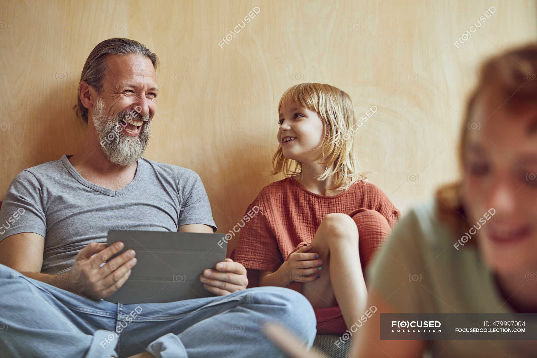 Cheerful father and daughter sitting against wall at home ...