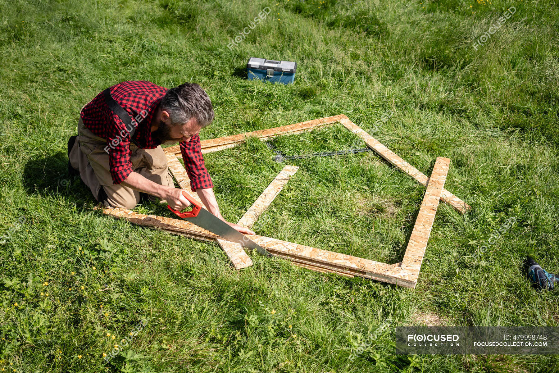 Carpenter man cutting plank with hand saw on grass during sunny day — outdoors, wood Stock