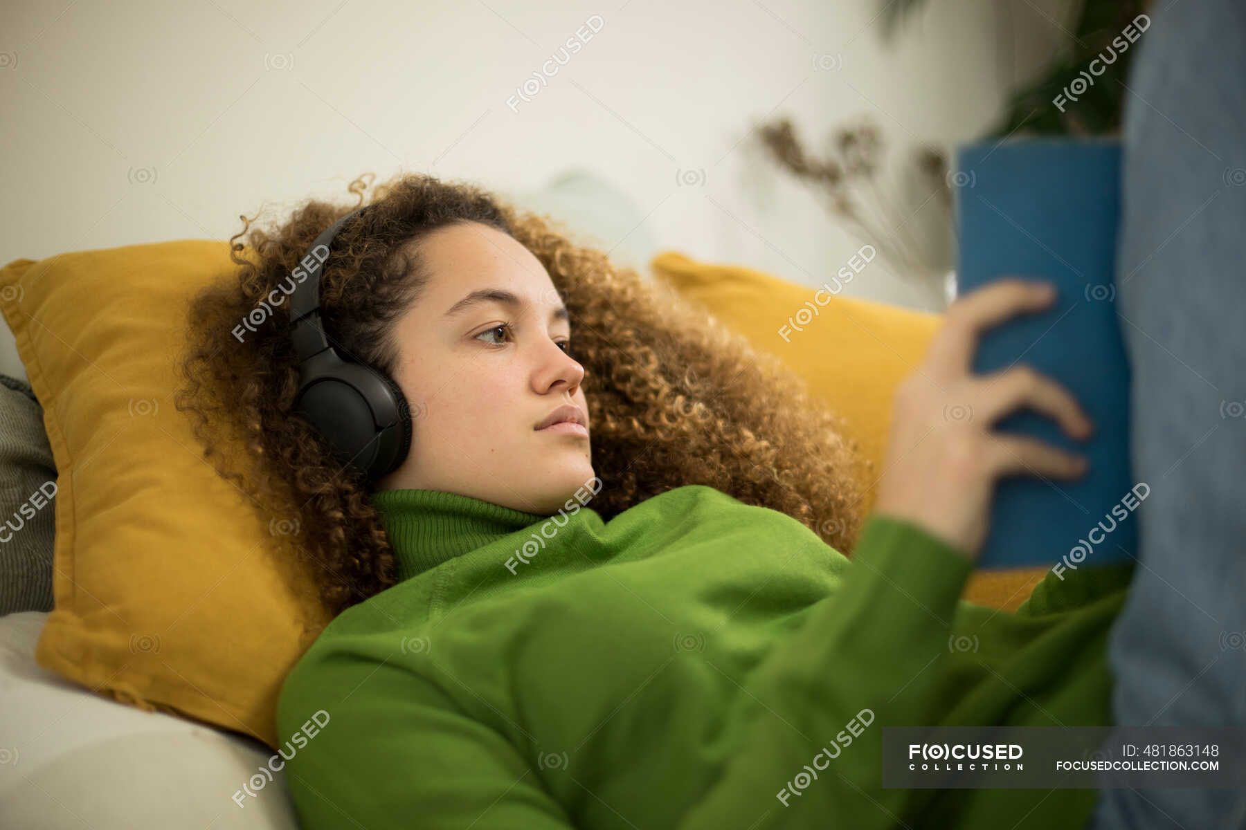 Молодая женщина с книгой лежит на кровати, слушая музыку в наушниках .