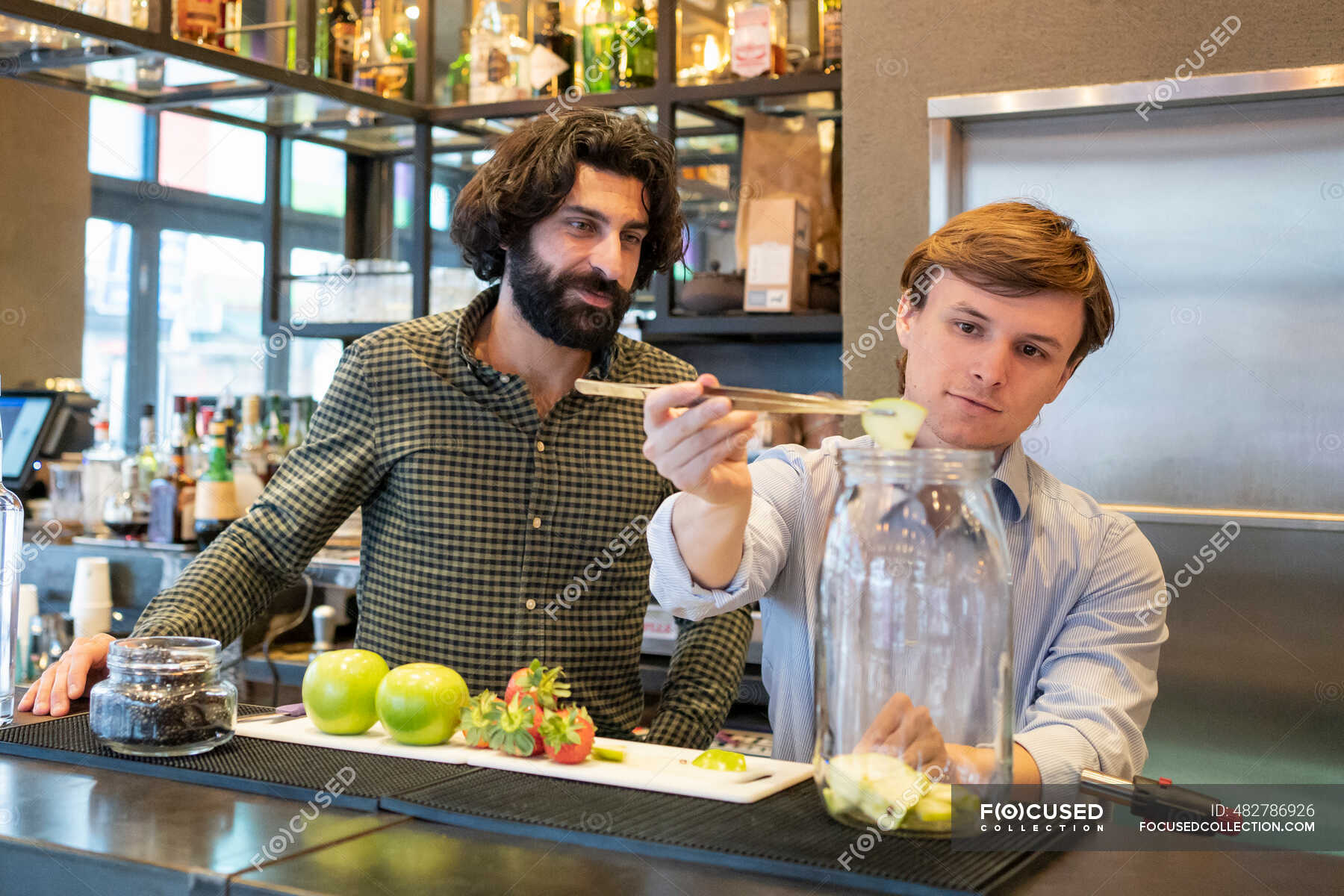 Male bartender looking at trainee putting apples in glass jar at bar