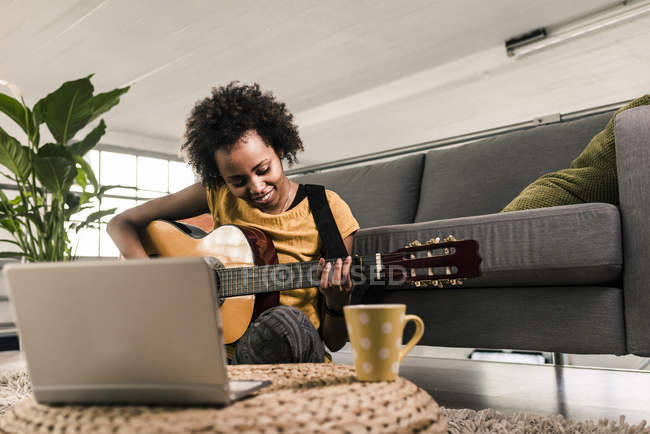 Sonriente joven sentada en casa con portátil y tocando la guitarra - foto de stock