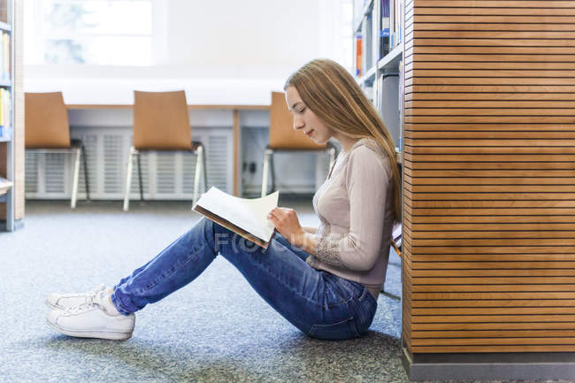 Teenage girl sitting on the floor in a library and reading book — Stock Photo