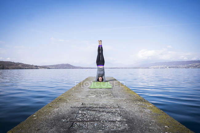 Femme pratiquant le yoga faisant un appui-tête sur une jetée près d'un lac — Photo de stock