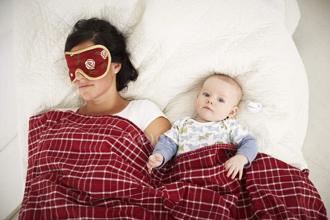 Mãe com máscara de sono e bebê acordado deitado na cama — Fotografia de Stock