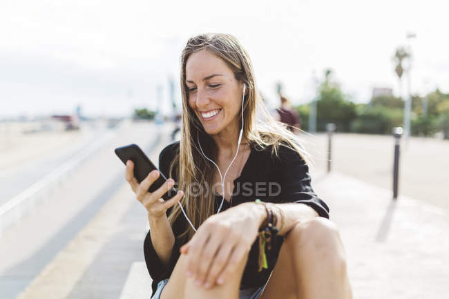 Lächelnde junge Frau mit Handy und Ohrhörern an der Uferpromenade — Stockfoto