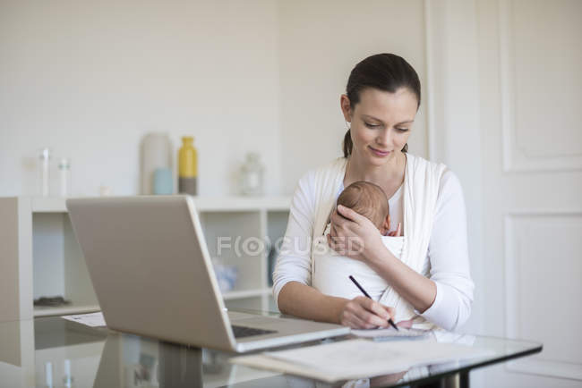 Freiberufliche Mutter mit Säugling im Tragetuch arbeitet zu Hause im Büro — Stockfoto