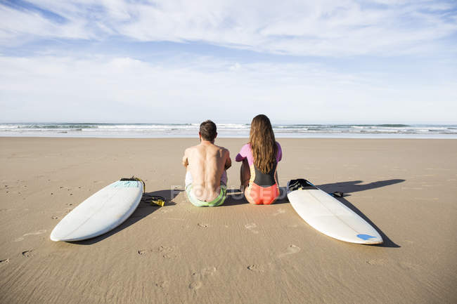 Rear view of surfers couple sitting at the beach with surfboards looking at se — Stock Photo