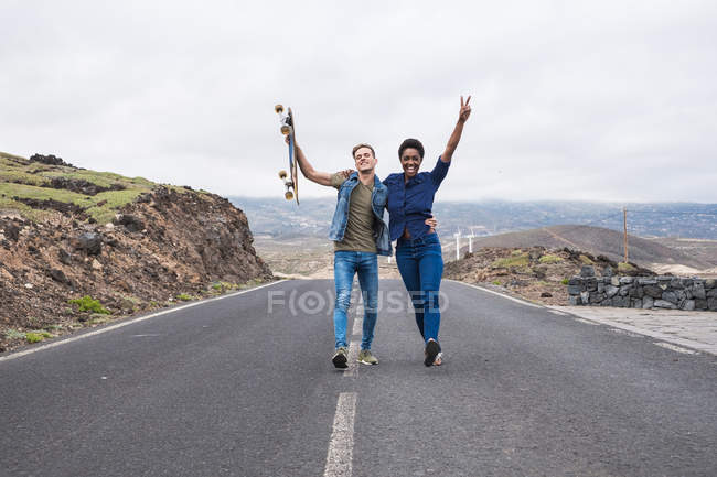 Attractive multicultural young couple with skateboard walking on road with raised arms — Stock Photo