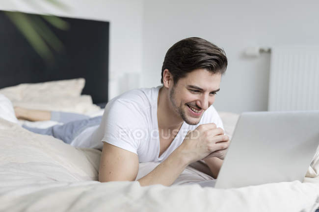 Uomo sorridente sdraiato sul letto e utilizzando il computer portatile — Foto stock