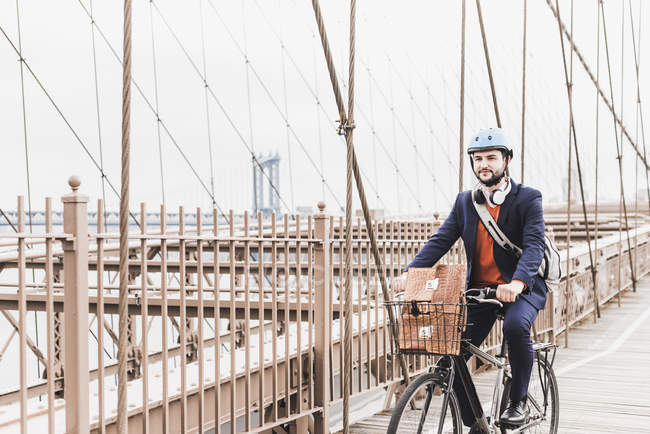 Estados Unidos, Nueva York, Brooklyn bridge, Hombre joven en casco en bicicleta con teléfono inteligente, paisaje urbano en el fondo - foto de stock