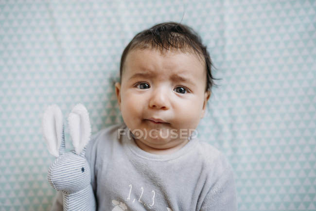 Bebê menina deitada no berço com rosto preocupado engraçado e segurando o coelho recheado — Fotografia de Stock